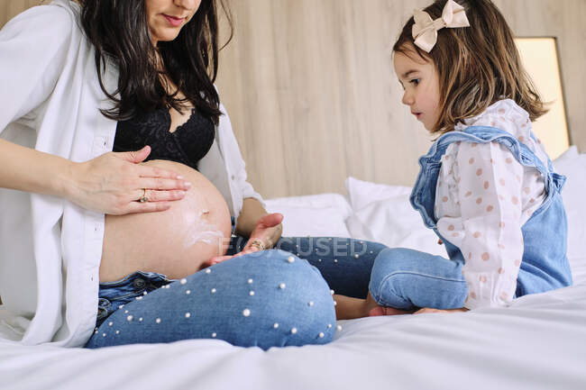 Girl looking at mother applying moisturizer on belly while sitting on bed — Stock Photo