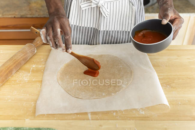 Hombre poniendo salsa de tomate en masa de pizza en la cocina en casa - foto de stock