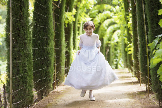 Cute girl in white communion dress running on footpath in green garden — Stock Photo