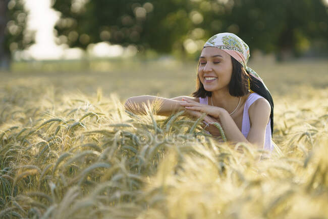 Улыбающаяся молодая женщина, стоящая с закрытыми глазами и наслаждающаяся пшеничным полем — стоковое фото