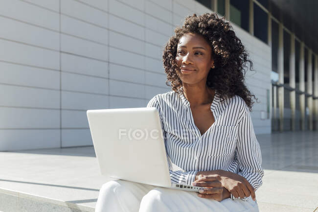 Femme d'affaires réfléchie regardant loin tout en étant assis avec ordinateur portable à l'extérieur de l'immeuble de bureaux — Photo de stock