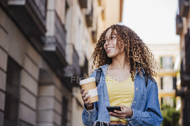 Bella donna guardando lontano, mentre tenendo tazza riutilizzabile e smart phone in città — Foto stock