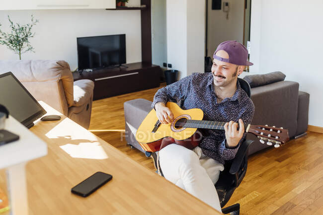 Guitariste masculin souriant portant une casquette jouant de la guitare à la maison — Photo de stock