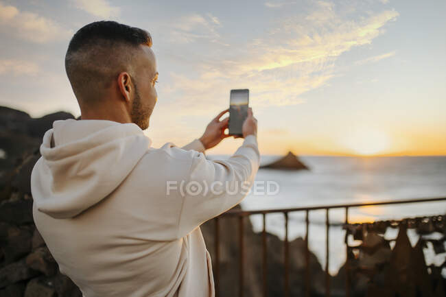 Uomo scattare foto di alba sul mare attraverso il telefono cellulare mentre in piedi sul ponte — Foto stock