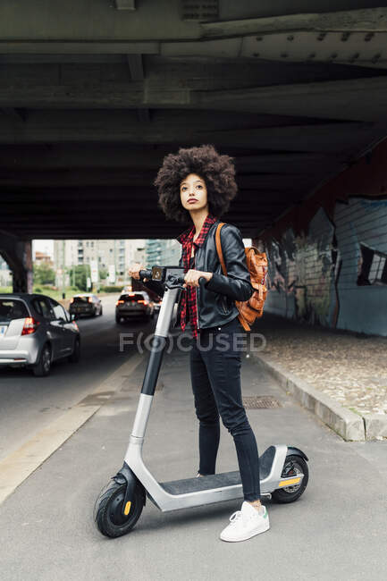 Молодая женщина стоит с электрическим самокатом на дороге под мостом — стоковое фото