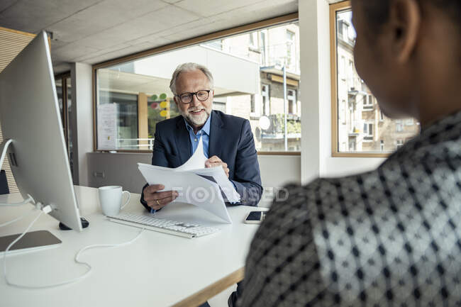 Sonriendo documentos profesionales masculinos de lectura delante de la computadora en la oficina - foto de stock