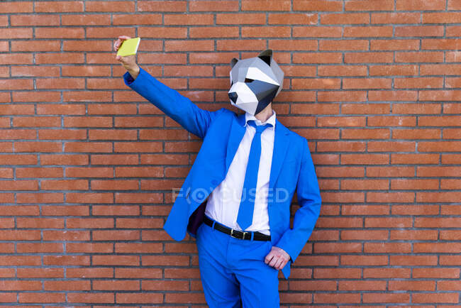 Hombre vistiendo traje azul vibrante y máscara de mapache tomando selfie teléfono inteligente contra la pared de ladrillo - foto de stock