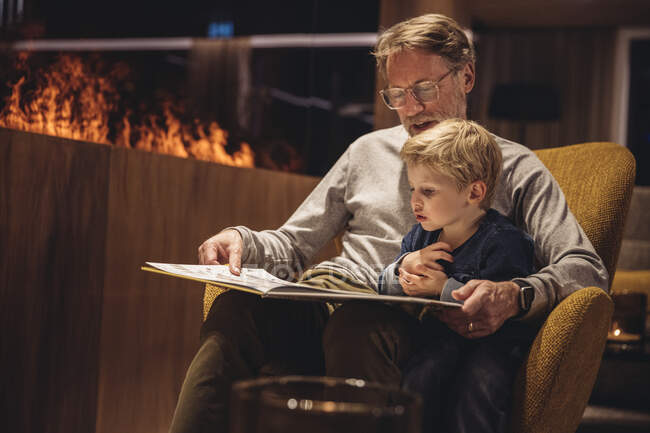 Vater mit Sohn liest Buch, während er im Wohnzimmer auf Stuhl sitzt — Stockfoto