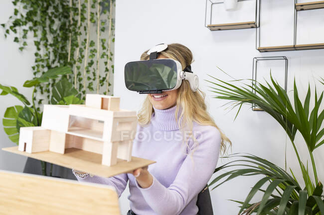 Sorridente professionista femminile che indossa un simulatore di realtà virtuale che tiene il modello di casa in ufficio — Foto stock