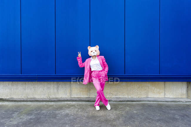 Женщина в ярко-розовом костюме и маске медведя делает знак мира перед синей стеной — стоковое фото