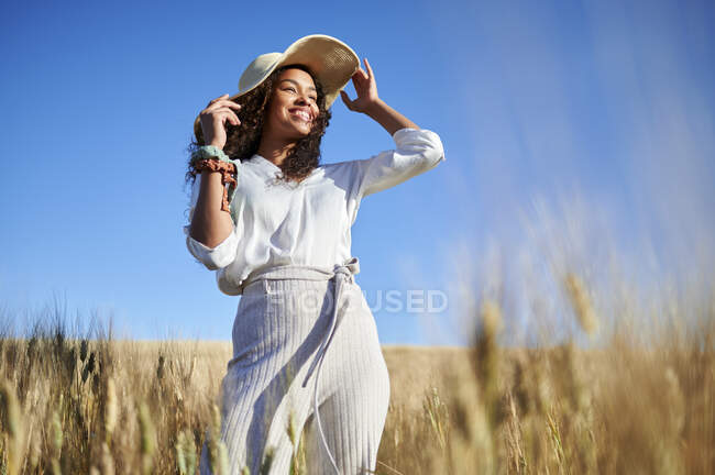 Улыбающаяся молодая женщина в шляпе смотрит в сторону, стоя на пшеничном поле — стоковое фото