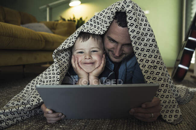 Улыбающиеся отец и сын с цифровой планшет лежал под одеялом в гостиной — стоковое фото