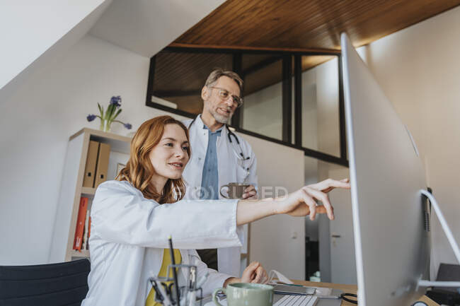 Médecin souriant pointant vers l'ordinateur tout en étant assis par un collègue au bureau — Photo de stock