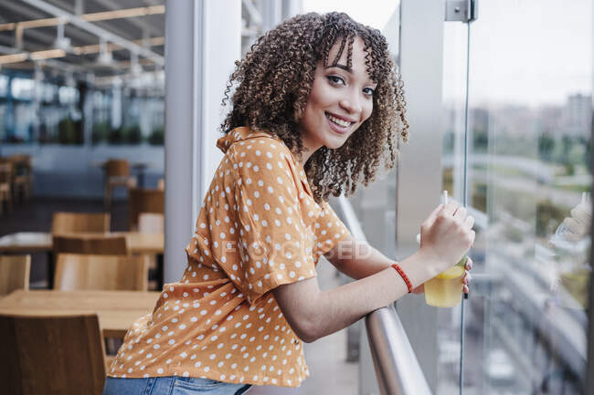 Sorridente giovane donna con tazza di tè usa e getta appoggiata su ringhiera al balcone in caffetteria — Foto stock