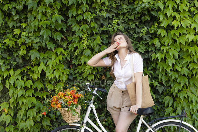 Young woman standing with bicycle blowing kiss in front of green ivy hedge — Stock Photo
