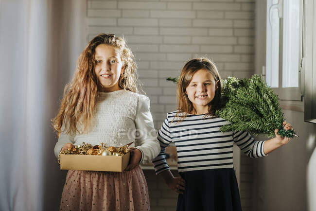 Lächelndes Mädchen mit Weihnachtsschmuckschachtel, Schwester mit Weihnachtsbaum auf der Schulter zu Hause — Stockfoto