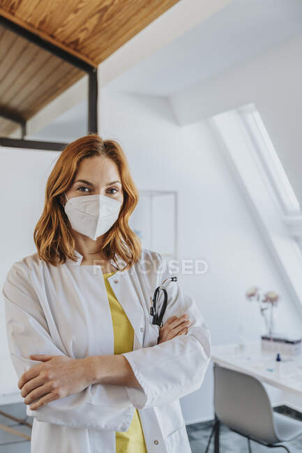 Doctora con mascarilla protectora de pie con los brazos cruzados en la clínica - foto de stock