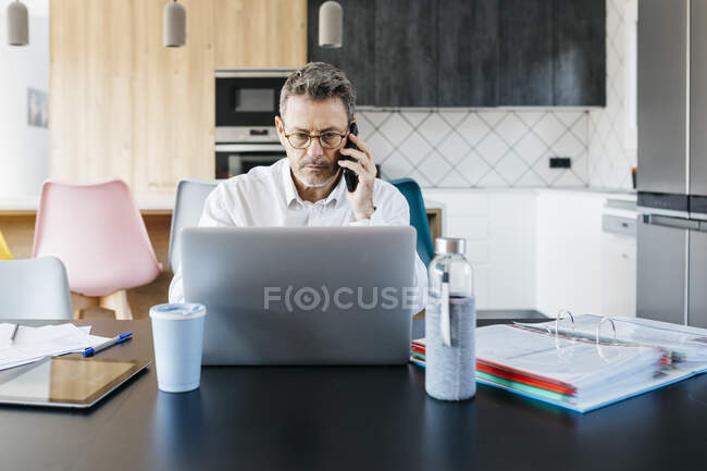 Серйозний чоловік-професіонал розмовляє на мобільному телефоні, працюючи на ноутбуці вдома — стокове фото