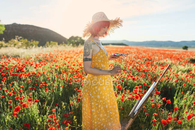 Artista feminina adulta média em amarelo sundress pintura no campo de papoula — Fotografia de Stock