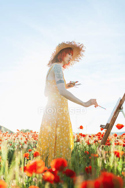 Mulher adulta média em chapéu de palha pintura na tela durante o dia ensolarado — Fotografia de Stock
