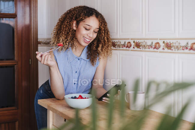 Beautiful woman using mobile phone while having fruit salad at domestic kitchen — Stock Photo
