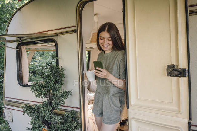 Sonriente mujer joven usando el teléfono inteligente de pie en la puerta del campista - foto de stock