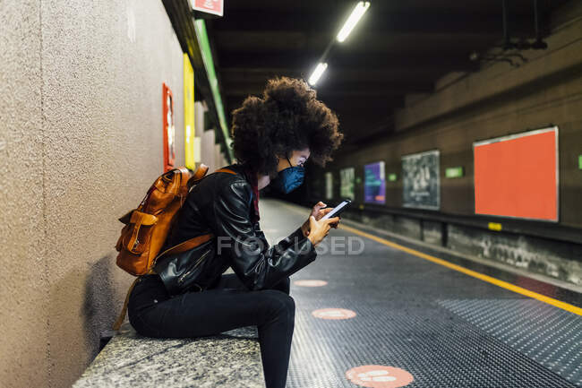 Женщина в защитной маске с помощью мобильного телефона, сидя на станции метро — стоковое фото