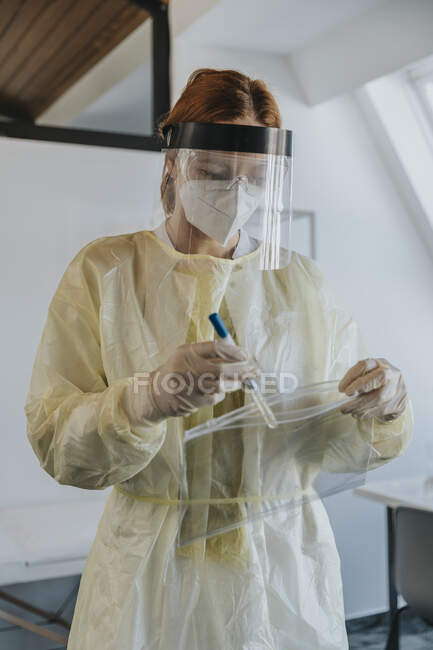 Doctora usando ropa de trabajo protectora poniendo muestra en bolsa de plástico mientras está de pie en la sala de examen - foto de stock