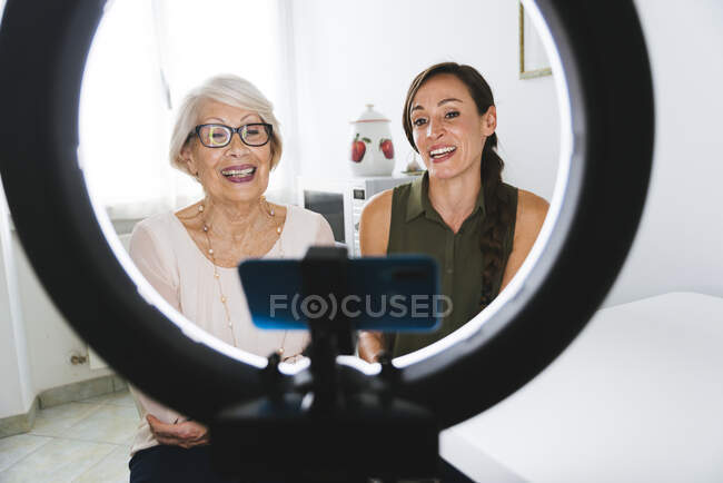Sonriendo a mediados de la mujer adulta vlogging con la abuela mientras se sienta en casa - foto de stock
