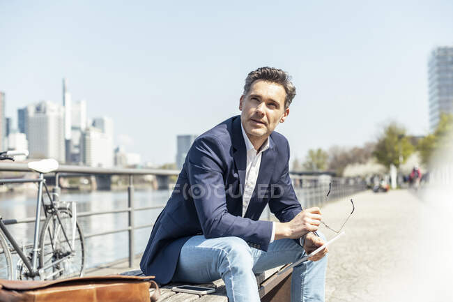 Задумчивый предприниматель смотрит в сторону, сидя на скамейке запасных в солнечный день. — стоковое фото