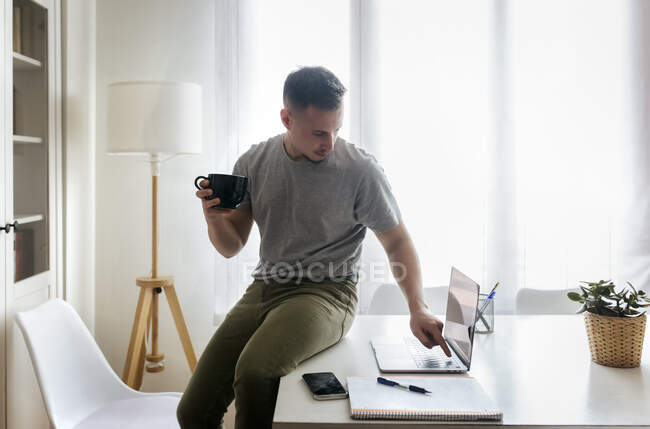 Homme d'affaires tenant tasse de café en utilisant un ordinateur portable tout en étant assis sur le bureau à la maison — Photo de stock