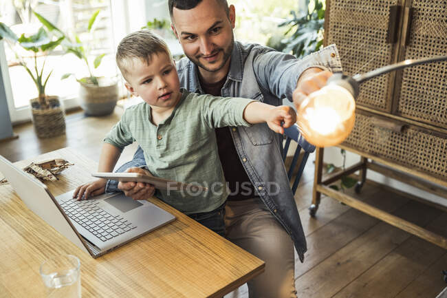 Hombre e hijo apuntando a la bombilla mientras están sentados con la tableta y el ordenador portátil en el hogar inteligente - foto de stock