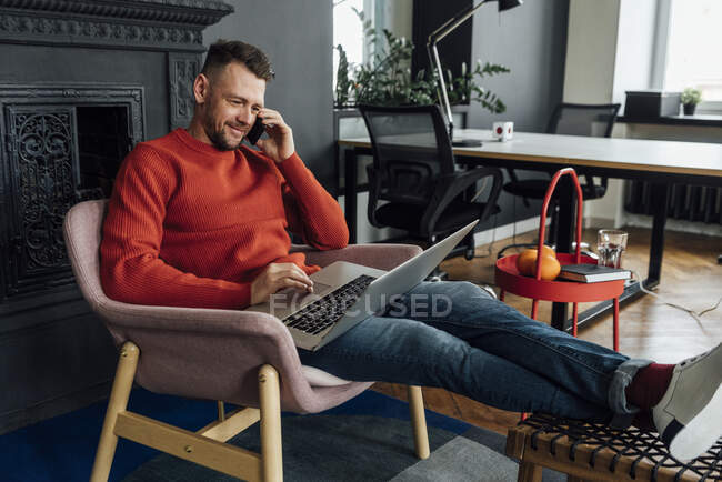 Empreendedor masculino usando laptop enquanto fala no telefone celular no escritório — Fotografia de Stock