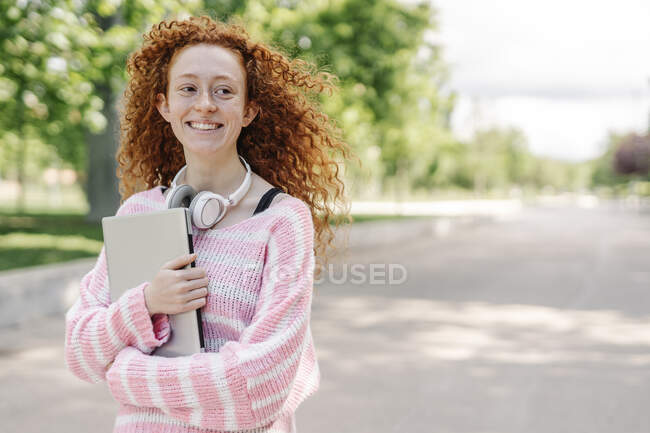 Happy young woman with curly hair carrying laptop in park — Stock Photo