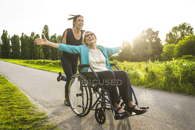 Bonne petite-fille poussant grand-mère assise en fauteuil roulant au parc — Photo de stock