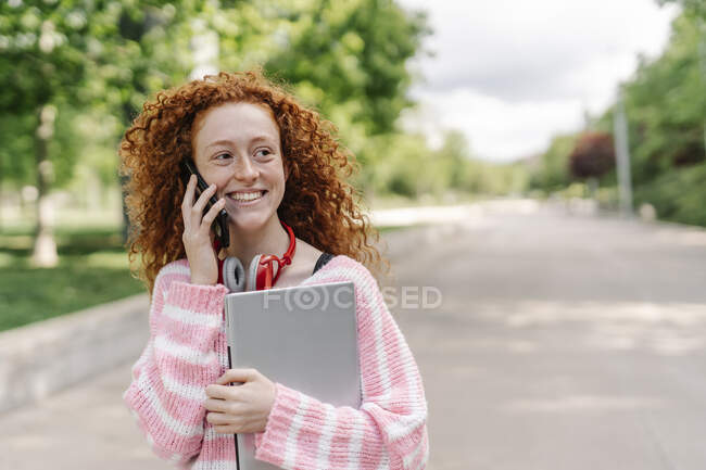 Joyeux rousse femme avec bouclé parler sur le téléphone intelligent au parc tout en regardant loin — Photo de stock