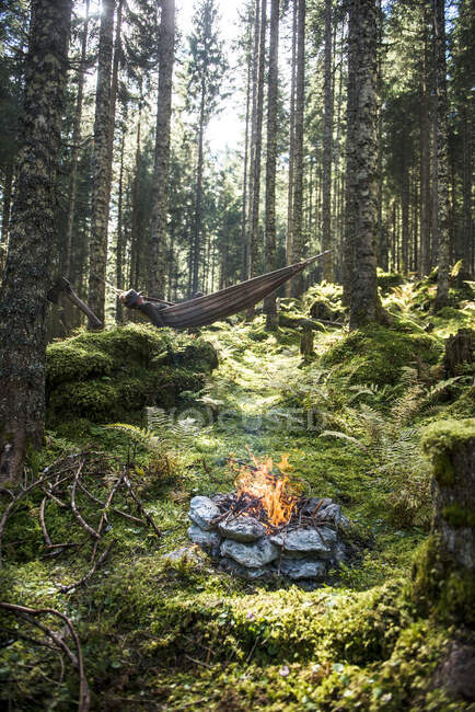 Fuoco di campo di pietra che brucia nella foresta con l'uomo sdraiato in amaca sullo sfondo — Foto stock
