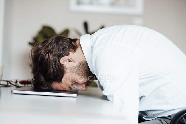 Hombre de negocios cansado apoyado en el ordenador portátil en la oficina - foto de stock