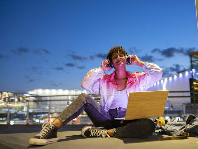 Mujer sonriente disfrutando de la música en los auriculares mientras se sienta al aire libre por la noche - foto de stock
