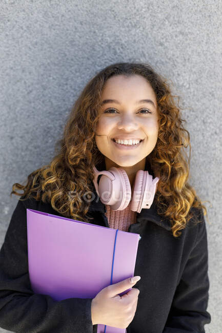 Mujer joven sonriente con auriculares sujetando el archivo contra la pared - foto de stock