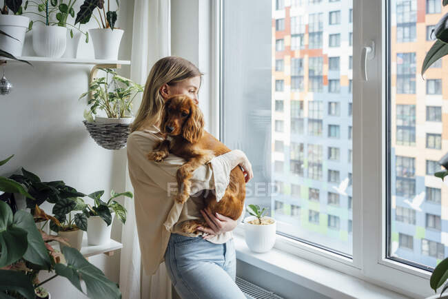 Blond woman with dog looking away while standing by window at home — Stock Photo