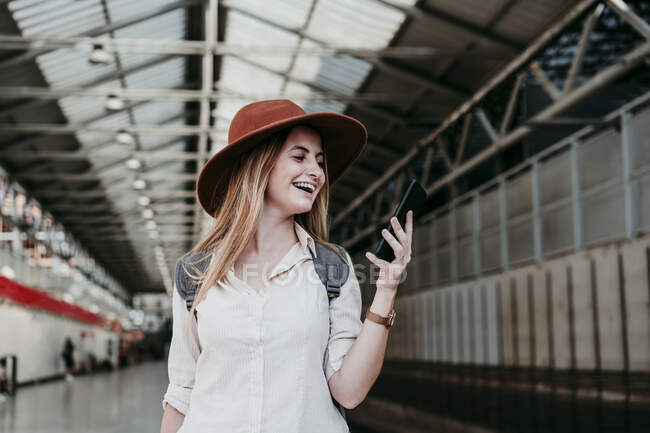 Веселая молодая женщина в шляпе с помощью смартфона на железнодорожной станции — стоковое фото