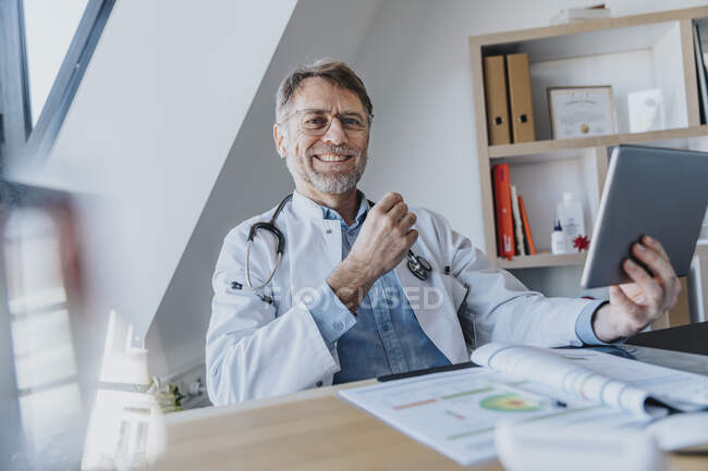 Medico maschio sorridente con tablet digitale seduto alla scrivania dello studio medico — Foto stock