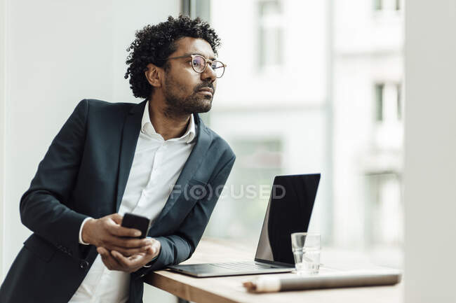 Entrepreneur masculin réfléchi avec téléphone intelligent appuyé sur le bureau tout en regardant par la fenêtre dans le bureau — Photo de stock