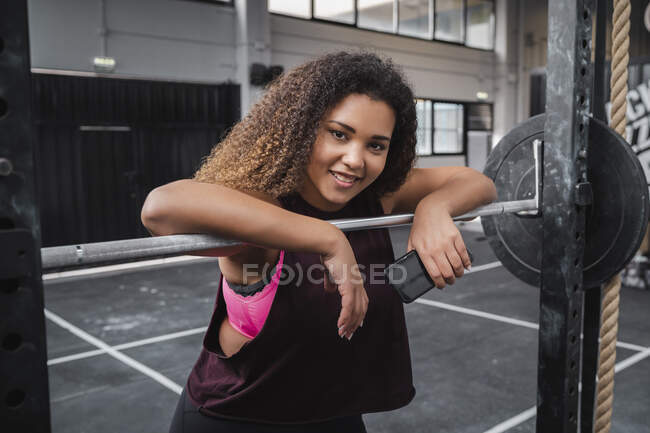 Smiling female athlete with mobile phone leaning on barbell at gym — Stock Photo