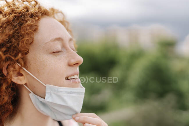Young woman with eyes closed removing protective face mask during pandemic — Stock Photo