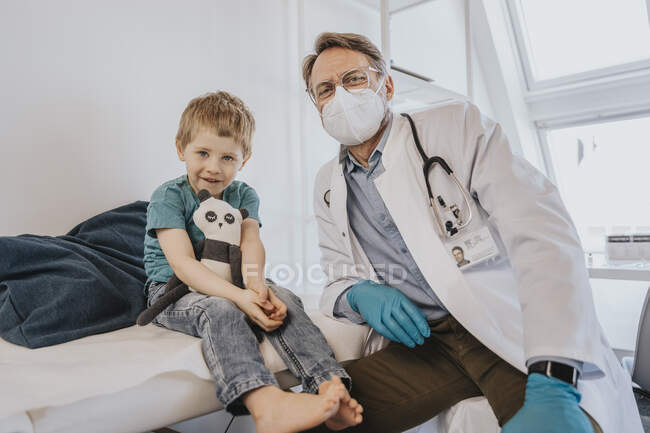 Médico com máscara protetora sentado por menino na sala de exame — Fotografia de Stock