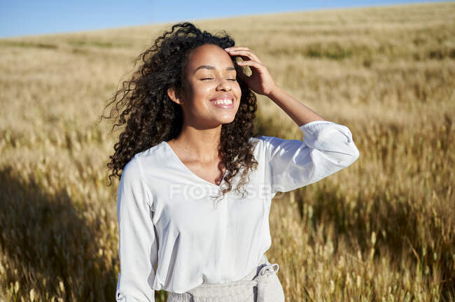 Femme aux cheveux bouclés souriante avec les yeux fermés debout dans le champ de blé par une journée ensoleillée — Photo de stock