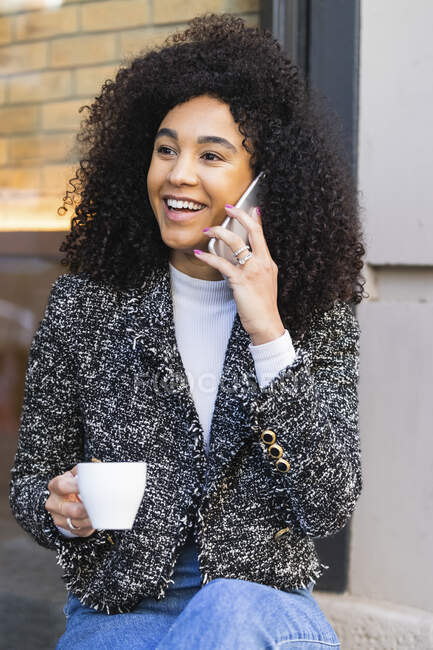 Mujer de pelo rizado sosteniendo taza de café mientras habla en el teléfono inteligente fuera de la barra - foto de stock