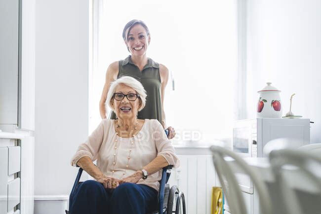 Посмішка серед дорослої жінки з бабусею - інвалідом сидить на стільці вдома на кухні. — стокове фото